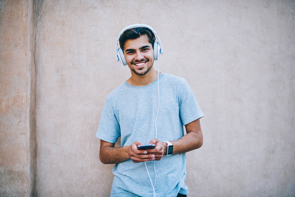 Portret van gelukkig hipster kerel glimlachend in de camera terwijl luisteren audioboek online in moderne hoofdtelefoon aangesloten op smartphone staande op promotionele achtergrond. Jonge man genieten van muziekspeler - Foto, afbeelding