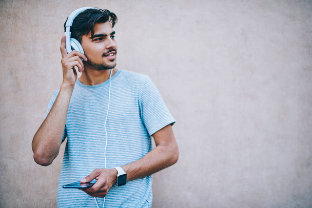 Positive hipster guy lover of music looking away while listening electronic songs from playlist in headphones connected to smartphone device standing on promotional background with copy space area - Fotó, kép