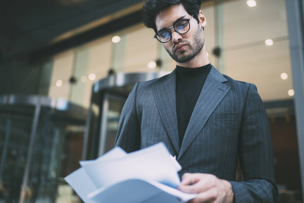 Pensive confident executive manager looking attentively at accounting reports and checking information standing outdoors.Successful entrepreneur reading financial paper documentation on street - 写真・画像