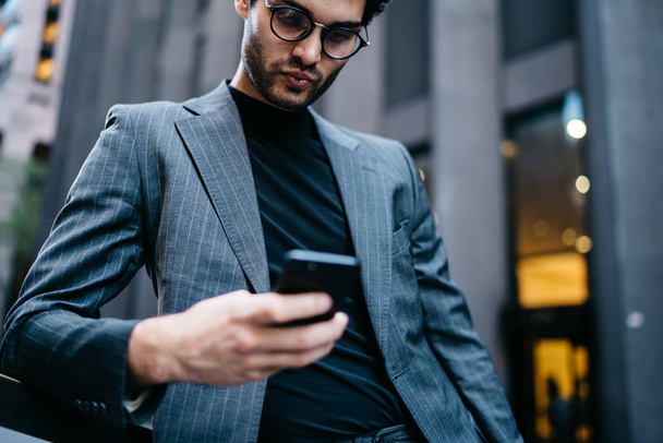 Bellow cropped image of confident formal dressed young entrepreneur reading incoming notification with finance balance on account on smartphone connected to 4G internet stnading near office building - Фото, изображение