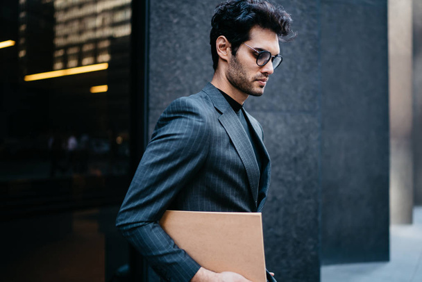 Confident young businessman dressed in stylish formal wear holding folder with financial documents in hand and strolling in urban setting near office building.Successful banker walking on street - 写真・画像