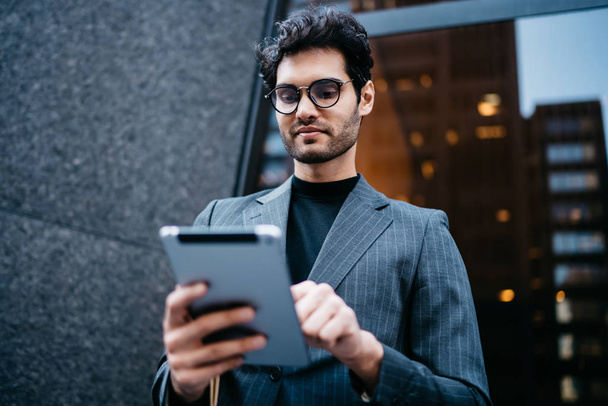 Below view of successful male entrepreneur dressed in elegant apparel downloading file with last business news on modern touch pad device connected to 4G internet standing near office building - Foto, Imagem