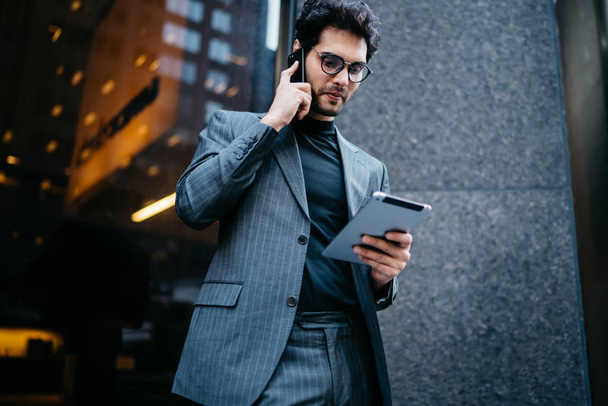 Prosperous entrepreneur talking on smartphone with business client and checking database on modern touch pad via 4G internet standing outdoors near office building.Successful lawyer talking on phone - Foto, Imagen