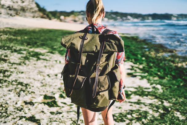 Young female tourist travelling with backpack and enjoying summer vacation and youth active lifestyle.Hiker with rucksack strolling outdoors on sea coastline in windy weather during trip - Photo, image