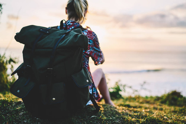 Cropped image of woman tourist enjoying summer vacation sitting on green grass coastline near to endless ocean.Back view of female traveler recreating with big rucksack during summer vacation - Foto, afbeelding