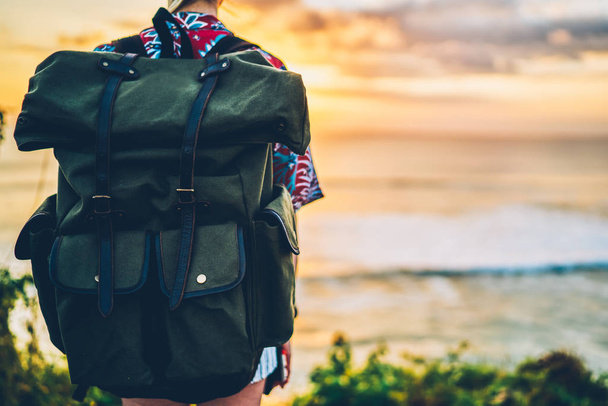 Cropped image of woman traveler carrying big green backpack while standing on coastline and observing sunset on horizon.Back view of hipster girl tourist spending summer vacation near to ocean - Photo, image