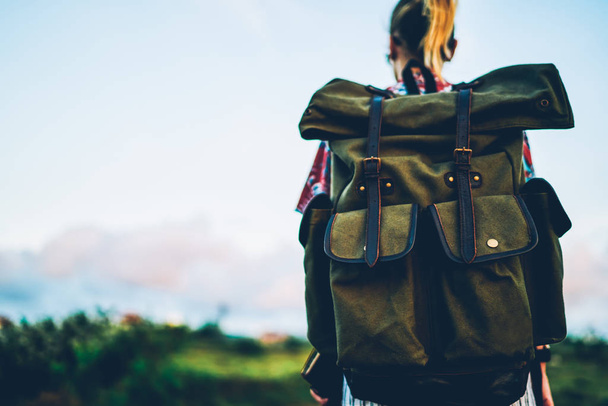 Cropped image of young woman tourist carrying big green useful for traveling backpack while walking across land at dusk.Back view of hipster girl with rucksack strolling during summer vacation - Foto, immagini