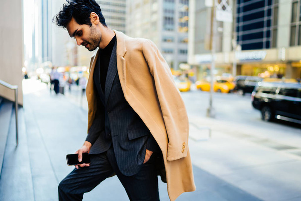 Thoughtful owner of company in stylish clothing walking holding mobile phone in New York financial district, trendy dressed young entrepreneur in elegant suit looking down spending time on stree - Foto, Imagen