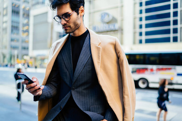 Pensive male entrepreneur in fashion clothes checking mail on smartphone and reading financial news, thoughtful businessman holding mobile phone while spending time on street browsing website online - Foto, immagini