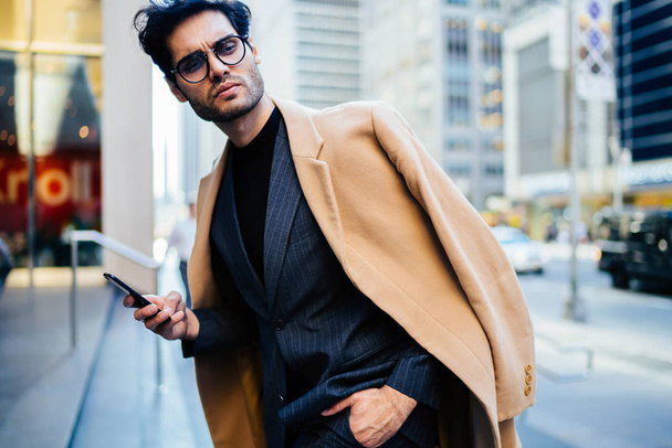 Vakava mies trendikkäissä vaatteissa ja silmälasien katselee pois seisoo kadulla älypuhelimen, mietteliäs komea liikemies ajatellut tehdä liiketoimen kautta hakemuksen matkapuhelimen ulkona
 - Valokuva, kuva