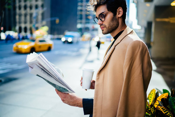 Pensive businessman in stylish outfit reading article in daily press starting day with morning coffee to go,confident executive manager concentrated on financial news in newspaper standing on stree - Fotó, kép