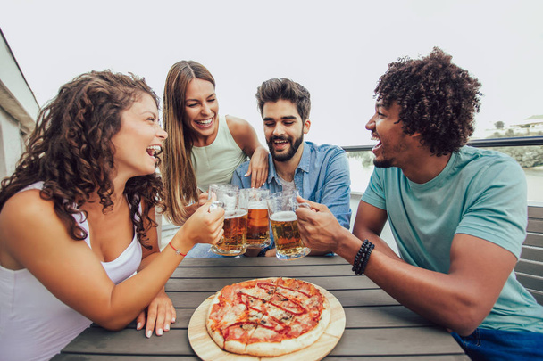 Grupo de jovens amigos alegres desfrutando de pizza e bebendo cerveja enquanto se sentam à mesa no telhado do prédio
 - Foto, Imagem
