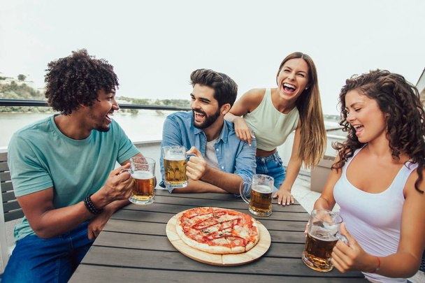 Gruppe junger fröhlicher Freunde genießt Pizza und trinkt Bier, während sie am Tisch auf dem Dach des Gebäudes sitzen - Foto, Bild