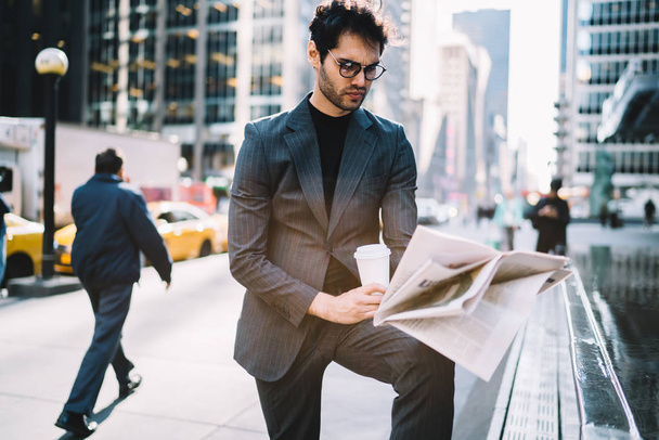 Concentrated prosperous owner in formal suit starting day with morning newspaper and coffee to go on Manhattan, serious executive male manager analyzing financial news in pres during break outdoor - Foto, Imagen