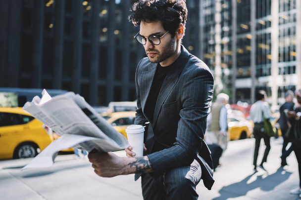 Handsome male professional in trendy formal outfit reading morning press with coffee to go in business district,confident man in elegant suit analyzing financial article in newspaper outdoors - Фото, изображение
