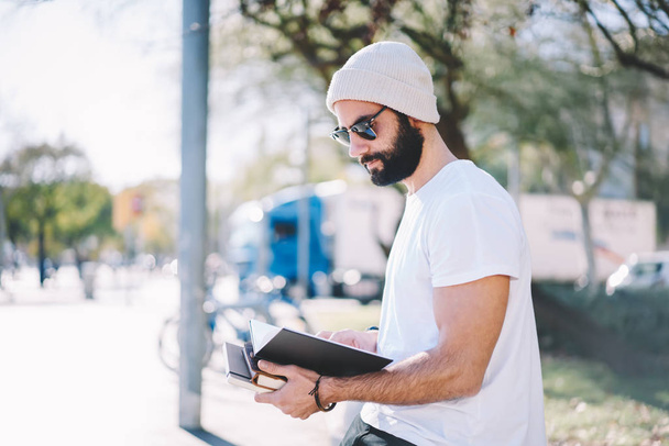 Nuori parrakas lahjakas mies opiskelija pukeutunut rento vaatteet lukeminen tietoa muistilehtiö viettäessään aikaa aurinkoinen street.Handsome fiksu hipster kaveri suunnittelee työaikataulu istuu ulkona
 - Valokuva, kuva