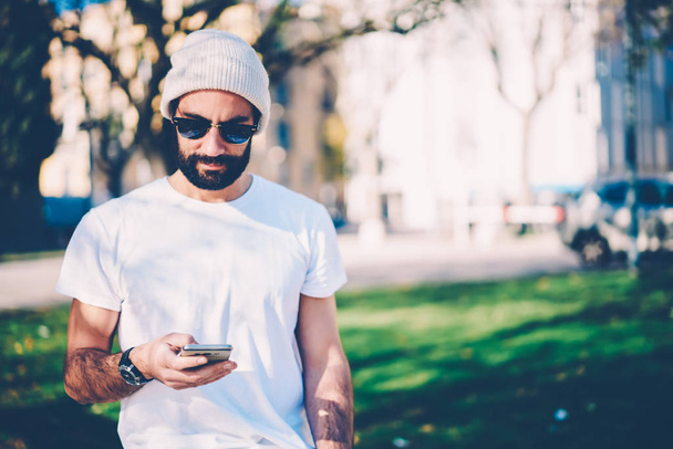 Handsome szakállas üzeneteket küld meg keresztül alkalmazás a smartphone, miközben felüdítő utca elegáns férfi. Csípő srác, online foglalás, és a kifizetéskor banki szolgáltatás a mobil - Fotó, kép
