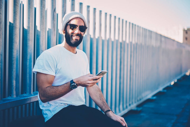 Half length portrait of cheerful man dressed in cool hat installing application from network via service on smartphone.Smiling male person looking at camera during sitting at urban setting with device - Zdjęcie, obraz