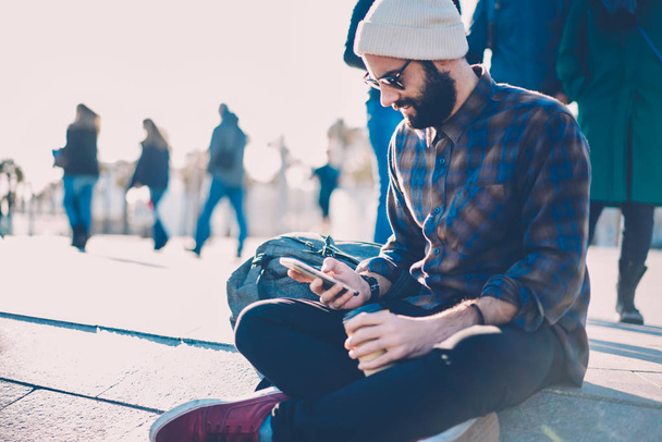 Bearded hipster guy dressed in casual shirt and stylish sunglasses sending notification to friend in social networks on smartphone via 4G internet connection sitting outdoors in urban setting - Zdjęcie, obraz