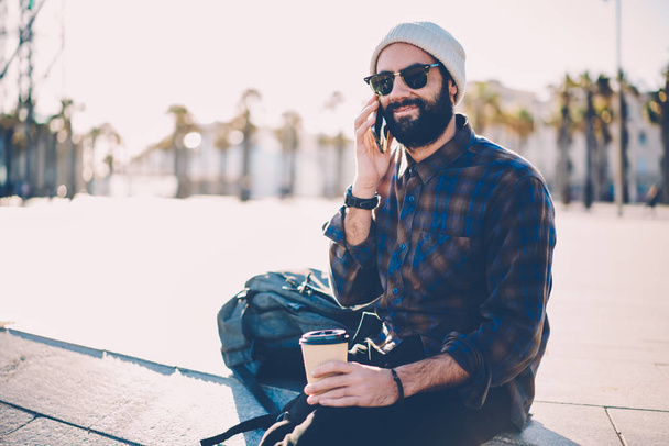 Positive stylish bearded man in sunglasses having mobile conversation on smartphone resting with coffee outdoors.Cheerful hipster guy in hat talking with friends on mobile phone enjoying beverage - Fotoğraf, Görsel