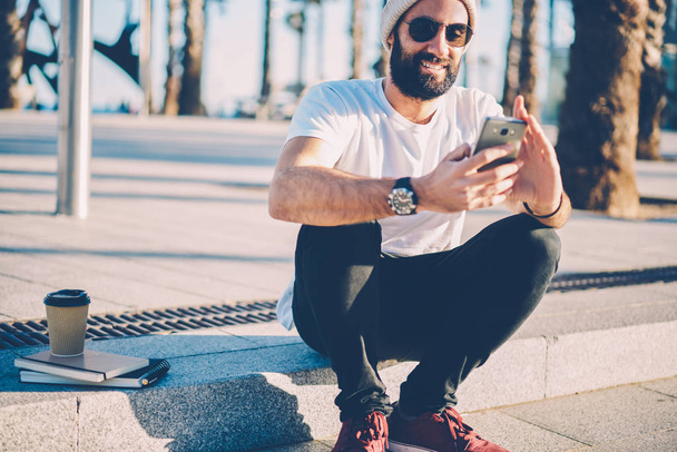 Випадковий одягнений бородатий студент в сонцезахисних окулярах читає новини на смартфоні, сидячи на відкритому повітрі в міських умовах.
 - Фото, зображення