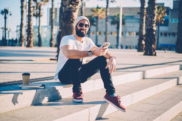 Stylish young bearded man in black sunglasses and trendy hat installing new mobile application on smartphone device via 4G internet connection spending leisure time outdoors sittting on stairs - Fotografie, Obrázek