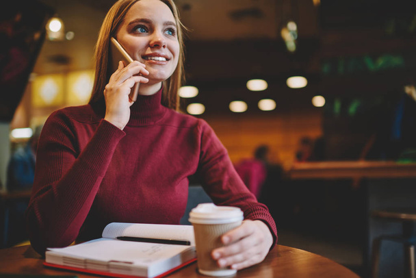 Cropped image of charming smiling blonde woman dressed in casual clothing having mobile conversation with friends in coffee shop.Young cute happy hipster girl talking on modern smartphone indoors - Photo, Image