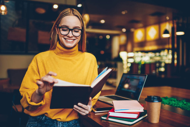 Charming emotional talented female student dressed in casual clothes sincerely laughing while spending time in coffee shop.Beautiful successful hipster girl reading information from notepad indoors - Фото, изображение