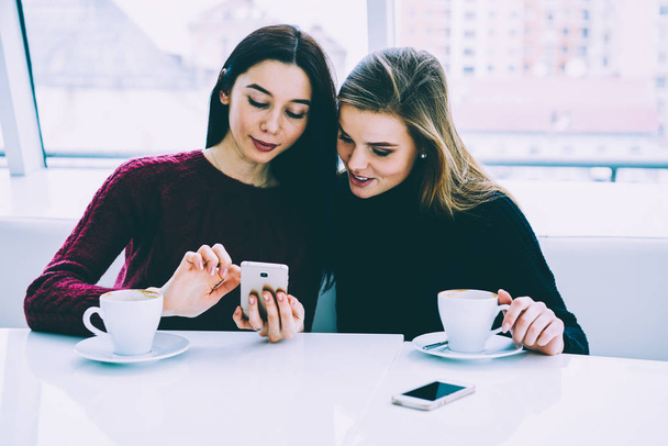 Positive beautiful friends dressed in trendy clothing reading funny news in website on cellular connected to 4G internet.Gorgeous females spending free time with modern gadget and tasty coffee - 写真・画像