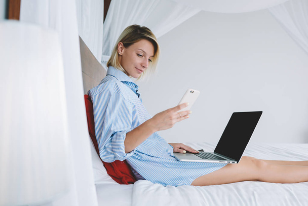 Positive young woman checking mail on smartphone in morning lying in bed of hotel room with laptop computer, female freelancer working remotely during weekend sending text message via cellular - Zdjęcie, obraz