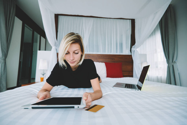 Young woman lying on bed with digital tablet for choosing items in web store during leisure, pensive female student reading content and new posts in social networks relaxing at home on leisure - 写真・画像
