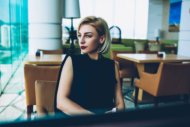 Gorgeous businesswoman with makeup and red lipstick dressed in elegant outfit looking away spending time in cafe interior, prosperous blonde female owner in formal wear thinking about future plans - Photo, image