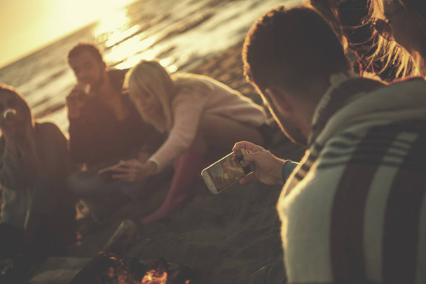 Happy Carefree Young Friends Having Fun And Drinking Beer By Bonefire On The Beach As The Sun Begins To Set filter - 写真・画像