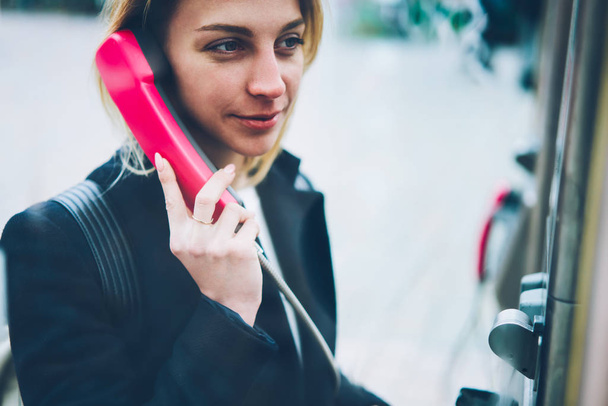 Positive blonde woman holding handset calling via stationery telephone on street, female tourist having conversation on public payphone operated with coins satisfied with international connection - Foto, afbeelding