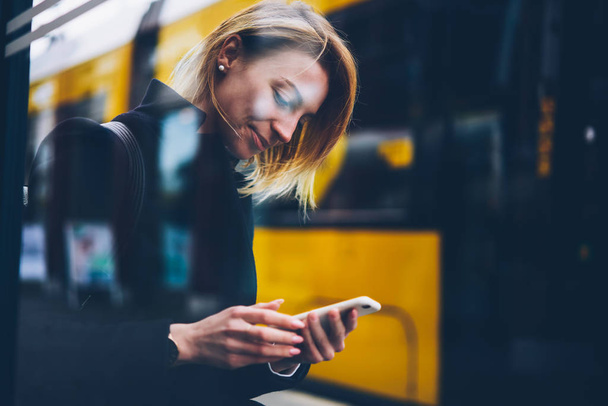 Positive blonde woman waiting for bus on stop browse application following traffic jams and ways in town, female tourist holding telephone satisfied with online news about transport standing on street - 写真・画像