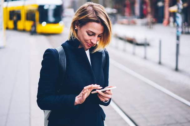 Positive blonde woman using smartphone for paying online for electric transport satisfied with city service, smiling female traveler checking routes of trams in cellphone app standing on street - 写真・画像
