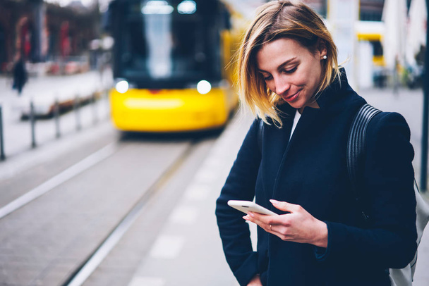 Smiling young woman checking table of city traffic online on web page using smartphone,positive hipster girl satisfied with app for paying for electric transport via cellphone waiting for tram - Фото, изображение