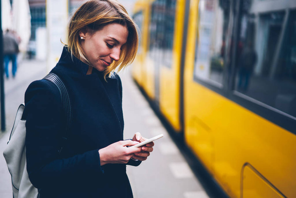 Blonde woman buying and paying for online transport ticket via banking application on smartphone satisfied with service, smiling female tourist following news about city traffic on cellphone - 写真・画像
