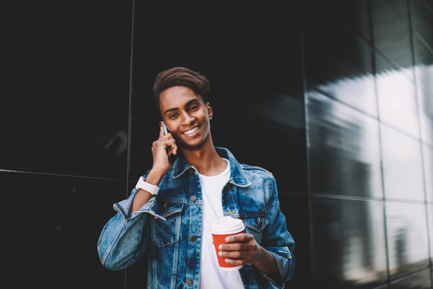 Half length portrait of positive hindu young guy dressed in stylish apparel talking on telephone and holding coffee to go in hands standing on black promotional background with copy space area - Fotó, kép