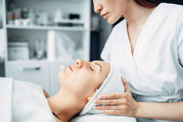 Beautician makes face massage to female patient, cosmetology clinic. Facial skincare, rejuvenation procedure in spa salon - Photo, Image