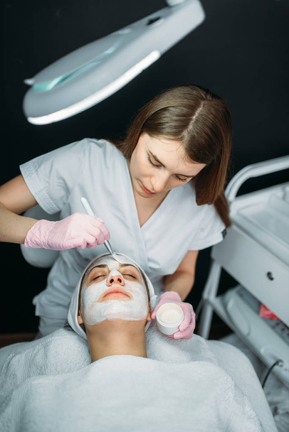 Female doctor holds cream and brush in hands, female patient on background, cosmetology clinic. Facial skincare, professional rejuvenation procedure in spa salon - Photo, Image