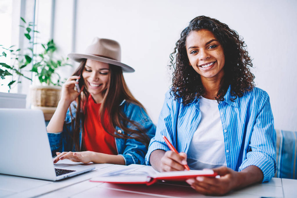 Portrait of cheerful female with curly hair looking at camera while writing in notepad spending time with friend, happy cheerful girl learning with colleague talking on phone about online courses - Foto, Bild