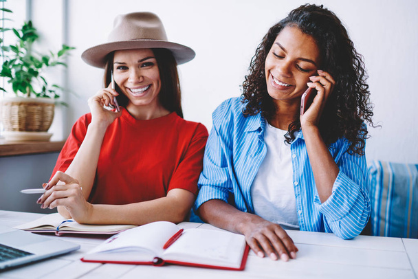 Cheerful female colleagues talking on smartphone during work break sitting in coworking office, happy trendy dressed hipster girls satisfied with god mobile connection and cheap tariffs making calls - Foto, Imagem