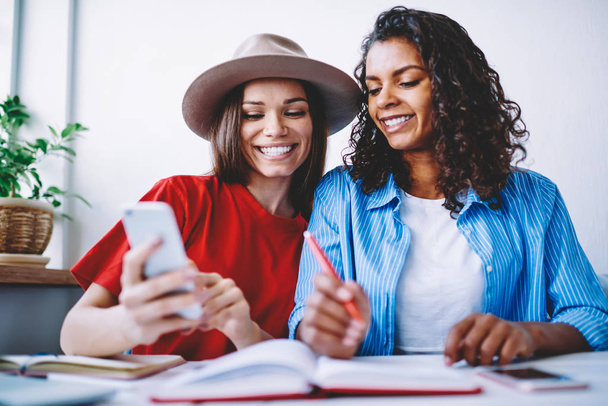 Cheerful female students having fun watching video from social networks while preparing for exams together, happy multiracial best friends viewing photos on mobile phone making bread during studying - Foto, Imagem