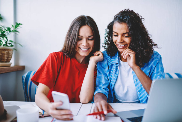 Happy female best friends excited with booking last tickets on web page browsing on smartphone,cheerful hipster girls watching funny videos on mobile making break during learning in coworking office - Фото, изображение