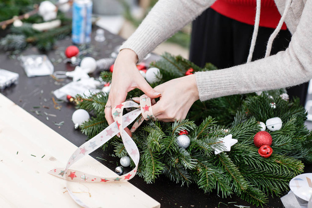 Woman tying a bow ribbon, decorated a Christmas wreath. Attaches toys and decor with glue gun. Hands close-up. Master class on making decorative ornaments. - Photo, Image