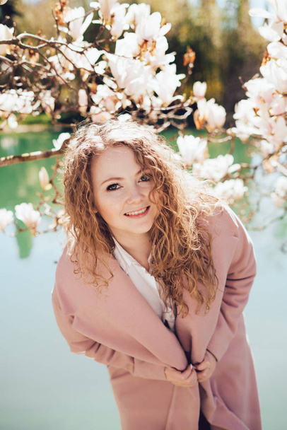 elegant curly woman in pink coat posing in park on background of flowers - 写真・画像