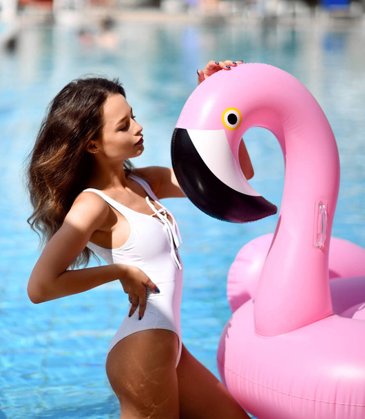 Young sexy pretty woman relaxing in a swimming pool leisure with giant inflatable giant pink flamingo float mattress in white bikini - Foto, imagen