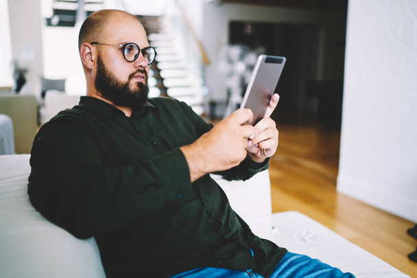 Concentrated casual dressed bearded man in eyewear reading e-book resting at home interior.Male blogger watching tutorial for installing app on tablet via wireless internet connection in apartment - 写真・画像
