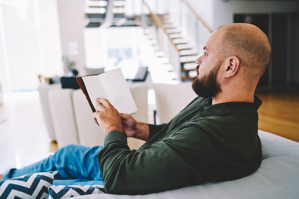 Concentrated bearded young man reading interesting literature book resting on comfortable sofa at home interior.Smart male with bestseller in hand enjoying recreation time in apartment - Fotoğraf, Görsel